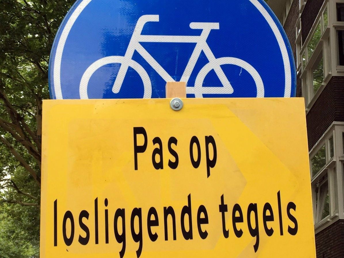 Waarschuwingsbord met de tekst: Pas op losliggende tegels fietsers en bromfietsers afstappen. Maar volgens een gestifte tekst dient men al sinds 2016 op te passen voor deze losliggende tegels.