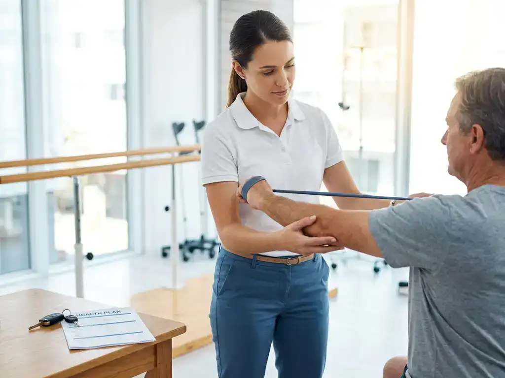 Revalidatietherapeut begeleidt patiënt bij armoefeningen in lichte therapieruimte, met parallelstangen en autosleutel op tafel op de voorgrond.